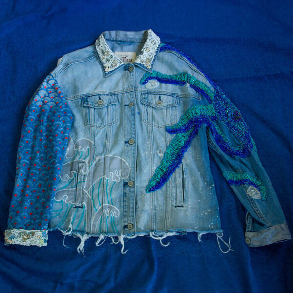 Front view of one of the Pulpis denim jackets with decorative embroidered white beading and buttons on the collar and cuffs. The right arm features a hand-painted wave texture, while the other arm showcases beaded tentacles in shades of blue. At the bottom of the jacket, there's a subtle ripped effect