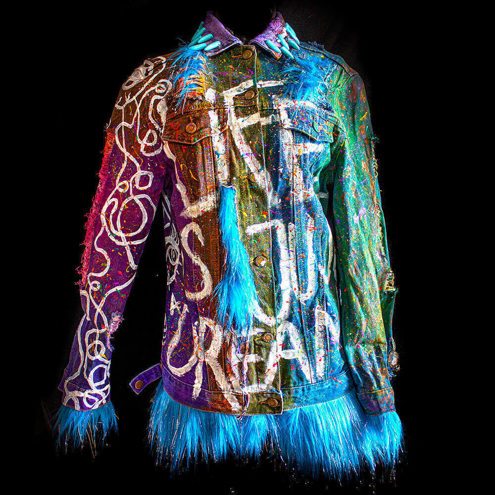 Front view of the Alien denim jacket, enhanced with furry textiles and adorned with hand-painted letters and various colored splatters on the fabric, achieved through a tie-dye process. Additionally, the jacket features turquoise spike beads added to the collar