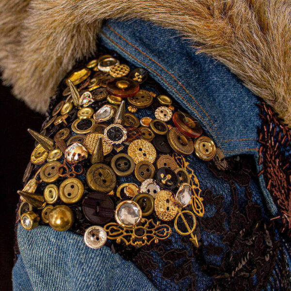 Close-up detail of the shoulder decorations on the denim furry hooded jacket. These decorations consist of buttons and conical metal spikes rivets in various sizes, featuring a combination of golden and copper colors. Additionally, black lace embellishes the area around the buttons on the shoulders