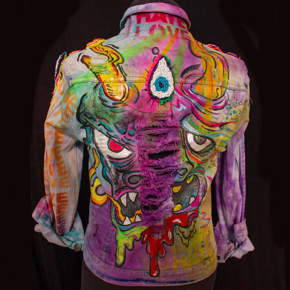 Back view of the Trippy Dragon denim jacket featuring neon and vibrant purple, red, and yellow paintings with a ripped texture at the back. The jacket showcases a hand-painted face of the dragon, as well as a removable eye-shaped beaded patch. The piece also incorporates text stencil texture, brick-like elements, and brilliant tie-dye colors