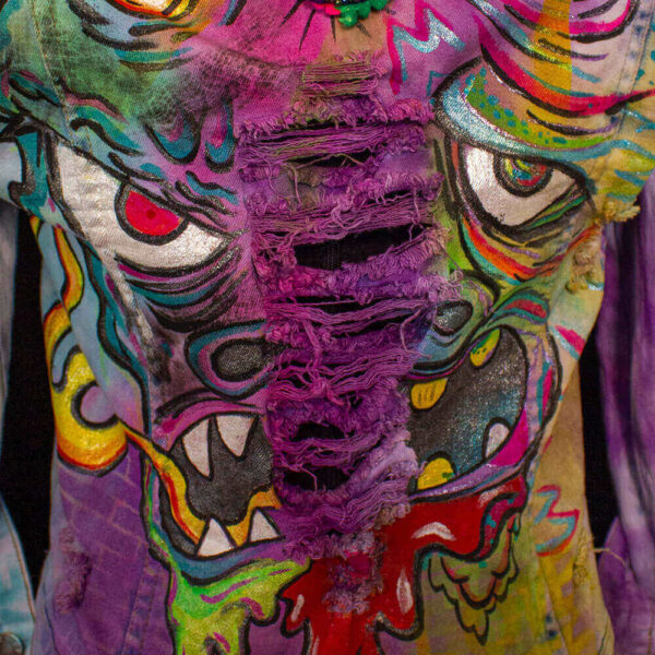 Close-up detail of the face of the Painted Trippy Dragon denim jacket. The dragon features red eyes, an open mouth with fangs, and green and red drool in the center of its face. The denim is ripped, showcasing neon purple, green, and yellow colors