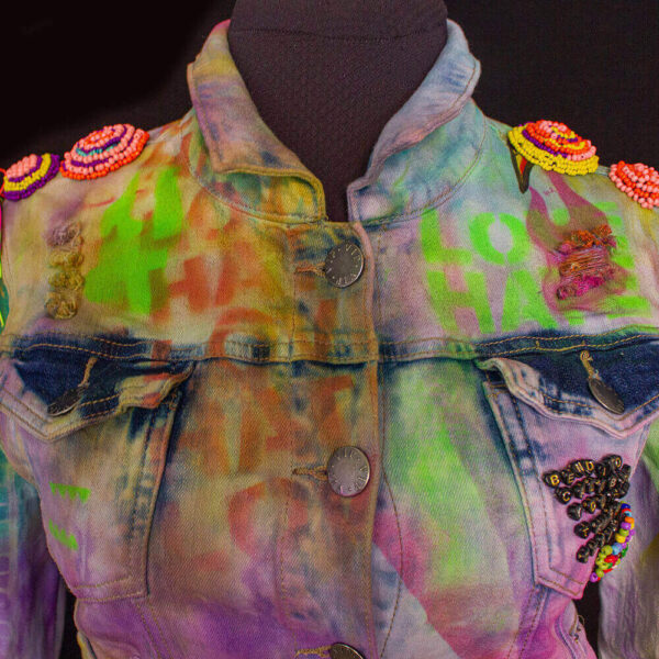 Close-up of the front of the Trippy Dragon denim jacket with tie-dye colors and a text stencil texture in vibrant neon shades. The shoulders feature removable beaded circles in psychedelic colors