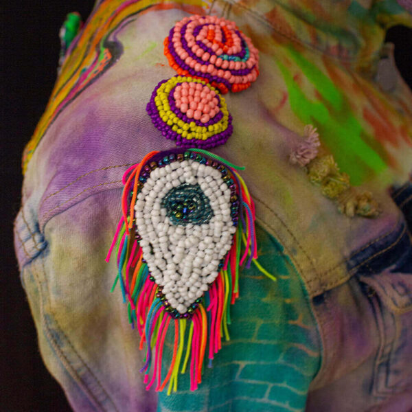 Close-up of the right shoulder of the Trippy Dragon denim jacket, featuring removable patches. There are two types of beaded patches: one is a drop-shaped eye with a white and black pupil, adorned with a colorful lanyard lariat strap around the border. The other patches are circle-shaped and display vibrant purple, yellow, and pink colors