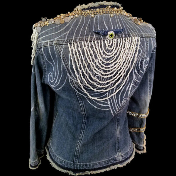 Back view of the Windy Jacket in denim with white bead decoration on the back. The jacket features a painted texture with white paint in lines, a removable beaded patch, white and transparent buttons on the shoulders, pearl and white beads, and golden chain decorations encircling the jacket's collar