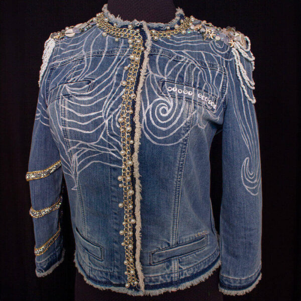 Front view of the Windy denim jacket with white lines painted and golden chains with white pearls along the collar and halfway down the jacket's opening. The shoulders feature a textured design with white beads, transparent and white buttons, and various other beads