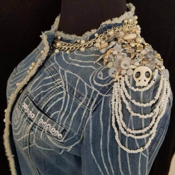 Left shoulder detail of the Windy Denim Jacket with white skull beads, transparent buttons, and white beads. It features a beaded cascade of white beads on the shoulder, along with white painted lines on the shoulder and front