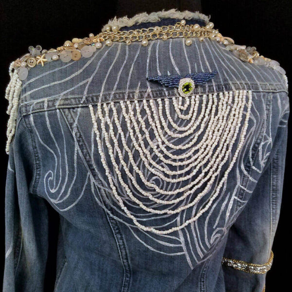 Back view detail of the Windy Denim Jacket with white painted lines and a cascade of white beads in a semi-circle at the back of the jacket. It also features a removable wing-shaped brooch at the center of the bead cascade