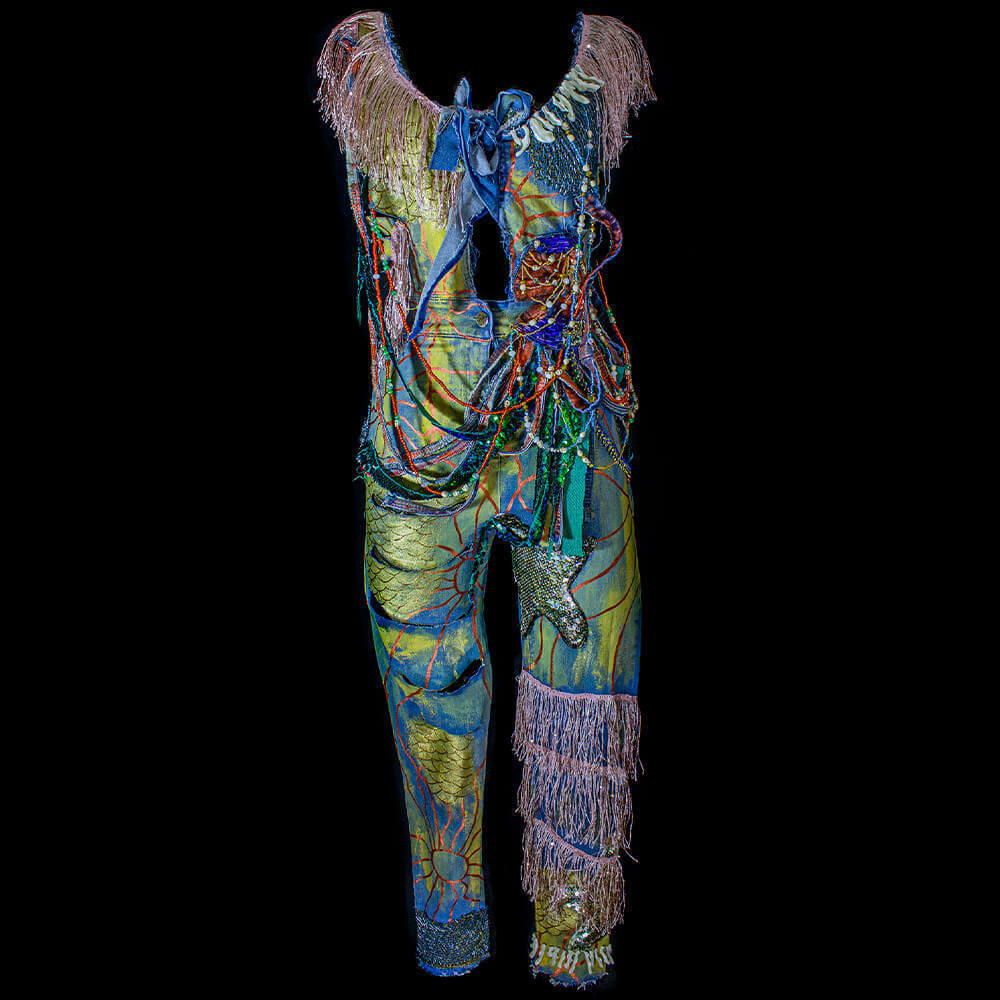 Front view of the Arrecife denim jumpsuit adorned with silver fringe trim, blue diamond net, and resin corals around the neckline. The jumpsuit is embellished with decorative long bugle beads, round colorful beads, and skull beads, forming a decorative net throughout the garment