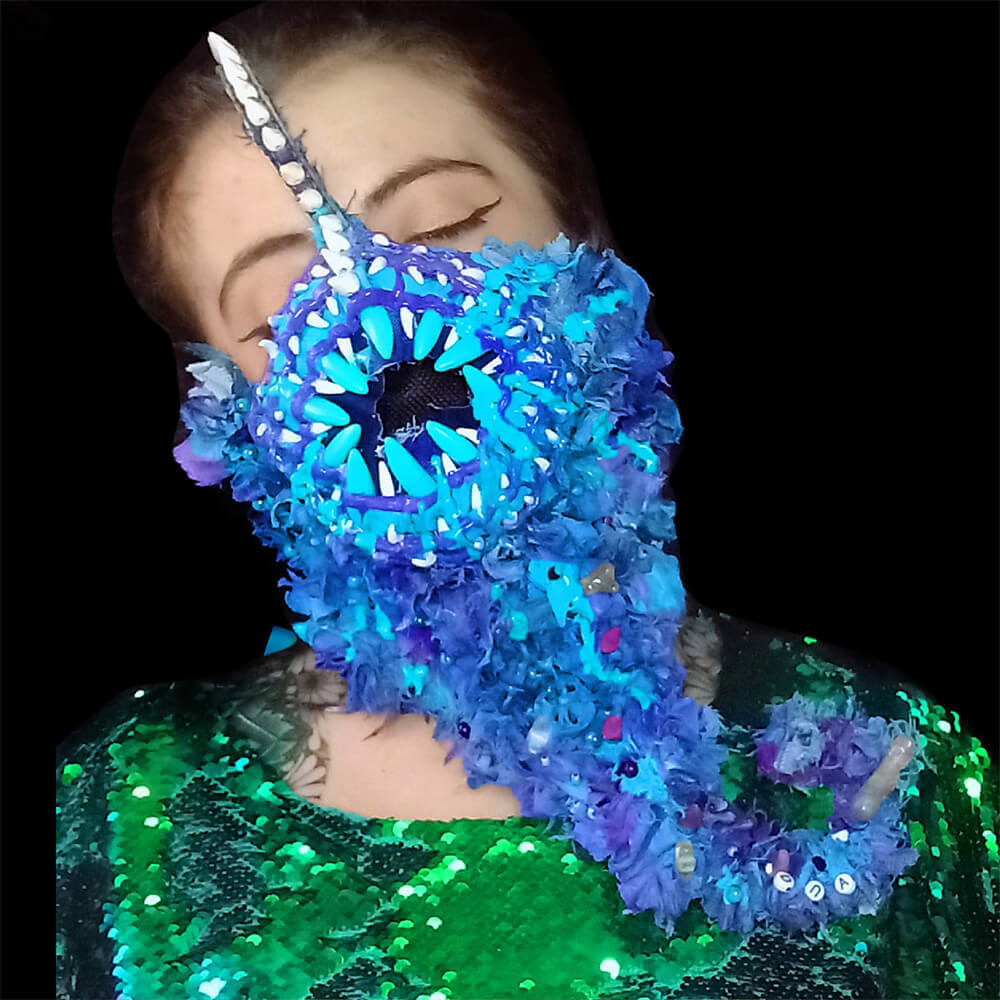 Front view of a model wearing the Barba Blue mask made with upcycled denim in purple and blue tones. Decorated with a striking mouth, glass beads, resin bones, 3D paint textures and fabric paint