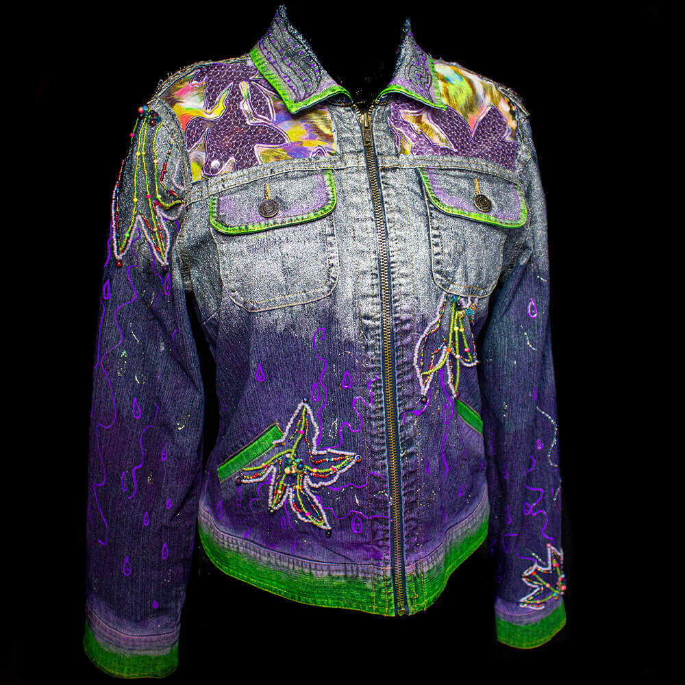 The Botanica Purple Denim Jacket features silver, purple, and green tones hand-painted onto the fabric, with added hand-painted lines. Additionally, the jacket boasts embroidery of colorful flowers with vibrant beads, as well as the incorporation of sequins and colorful patterns, adding to its unique and eye-catching design