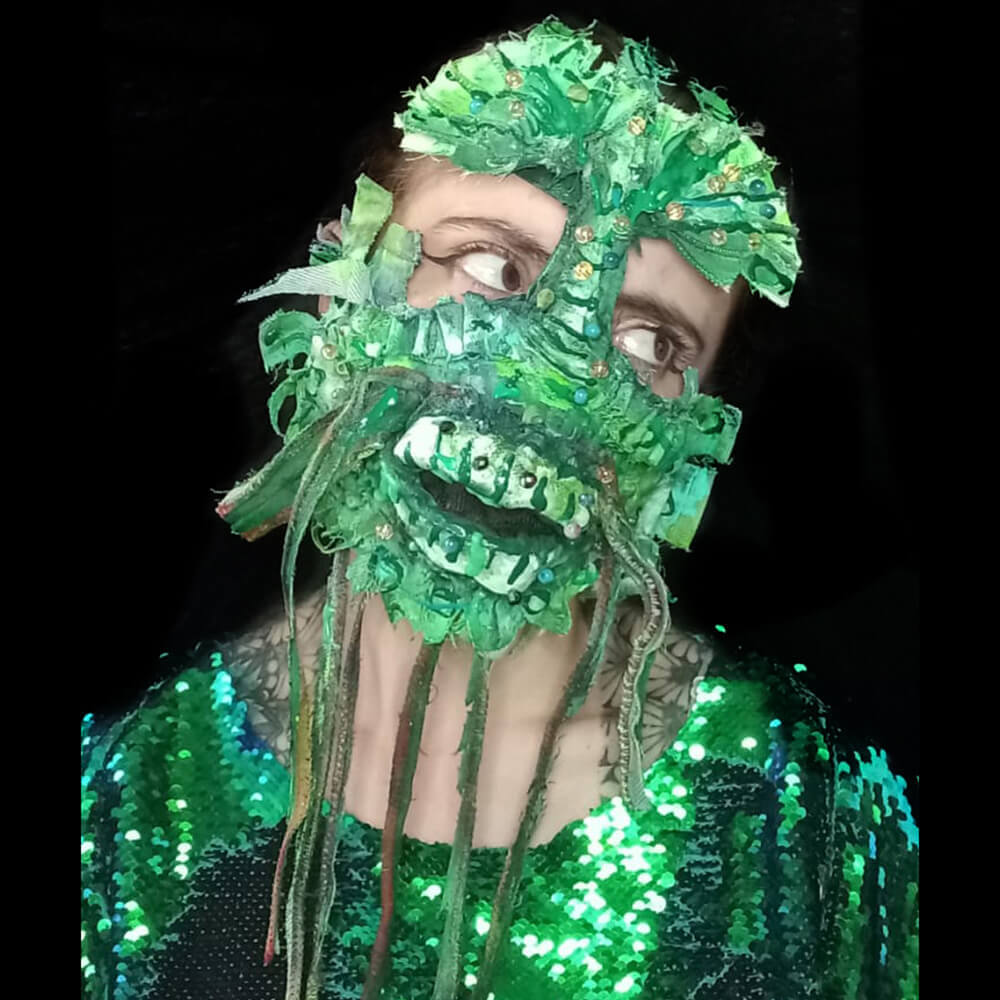 Front view of a model wearing the De la laguna upcycled denim mask decorated with green denim, golden & blue glass beads, 3D painting and fabric paint