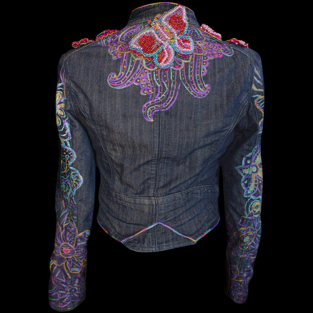 Back view of the personalized denim jacket - 'Eugenia Denim Jacket' decorated with an airplane and a butterfly beaded patch and with painted florals details with fabric paint and 3D paint