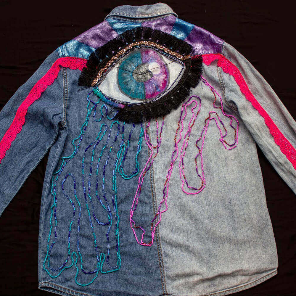 Back close view of the Eye Cry Denim Blouse a garment decorated with purple and blue fabric paint details below the collar details, featuring a captivating eye adorned with golden sequins and vibrant bead tears shape accents