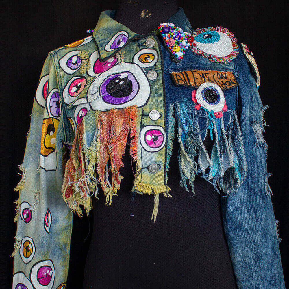 Front view of the Eyes Upcycled Denim Jacket decorated with removable bead eye shape patches in different sizes, details collar embroidered skulls, beads, fabric paint eyes drawings and ripped denim texture on edges