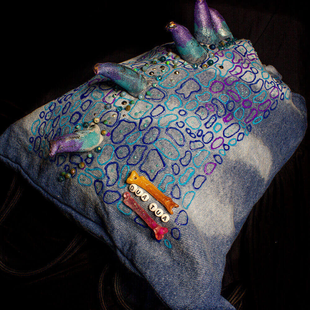 Top/right side general view of the Blue Iguana denim drawstring decorated with a crest made of denim scraps, embroidered decorative beads, 3D paint, resin bones accessories, letter beads, bleach denim effect and fabric paint