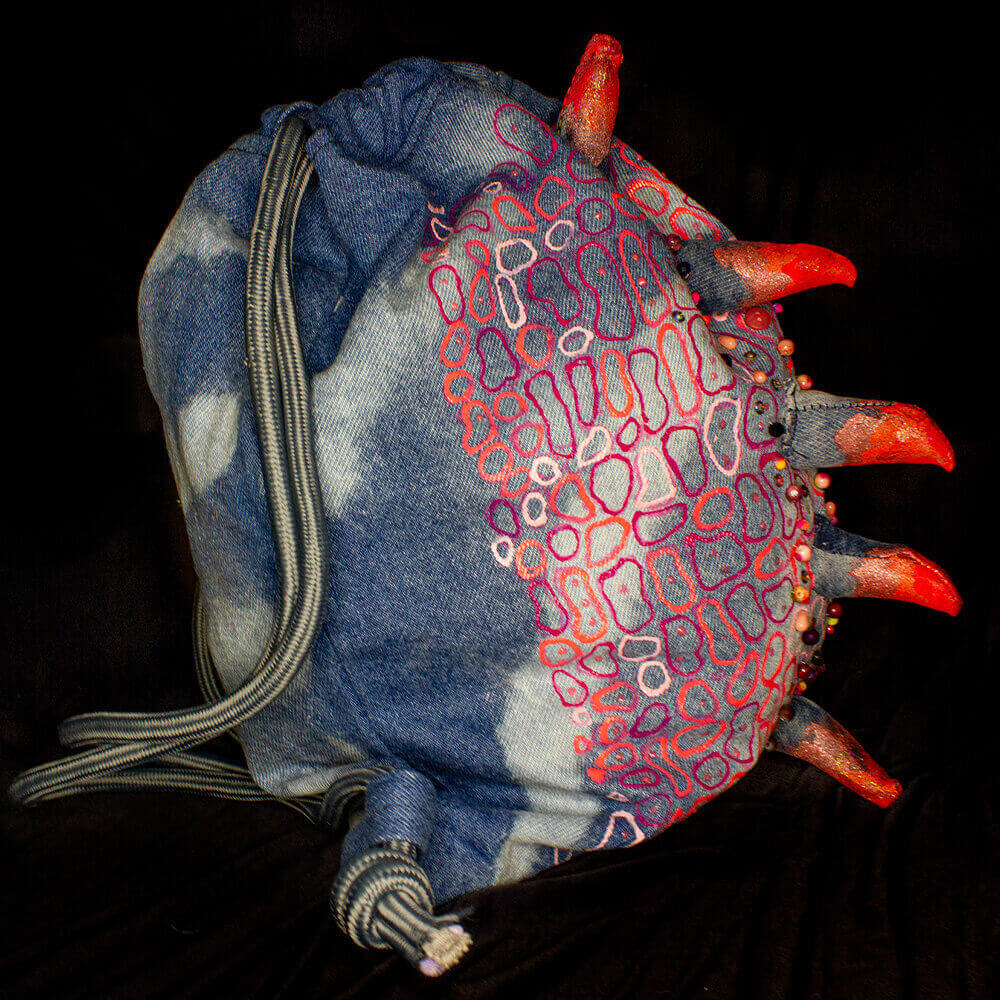 Right side view of the Pink Iguana denim drawstring decorated with a crest made of denim scraps, embroidered decorative beads, 3D paint, resin bones accessories, letter beads, bleach denim effect and fabric paint