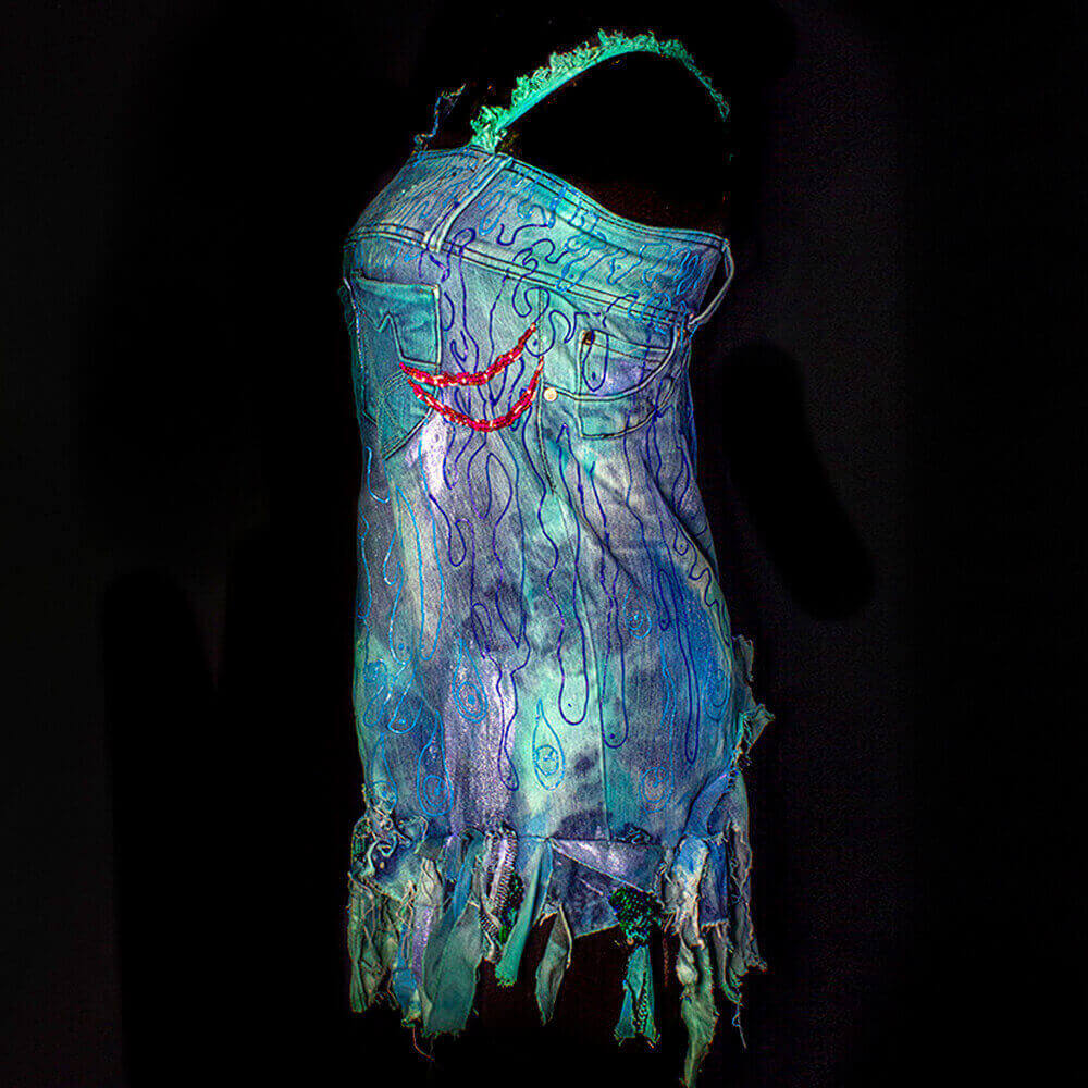 General left side view of the the Fluir denim dress made from an old pair of recycled denim jeans, decorated with denim scraps, hand embroidered beads as fish gills shape and 3D paint drawings, fabric paint and tie dye color process