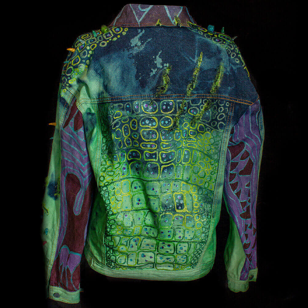 General back view of the Coco denim jack with a ripped mark on the back upper-right, a hand painted cocodrile-skin texture on the back, sleeves and collar, decorated with resin fangs and bird skulls on shoulders